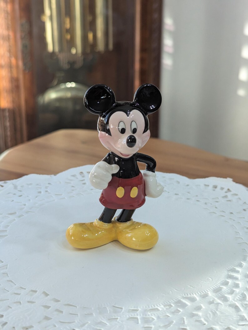 May include: A ceramic Mickey Mouse figurine. The figure has black ears, a black face, and a red shorts with two yellow buttons. The figurine is wearing yellow shoes and white gloves. The figurine is standing on a white paper doily.