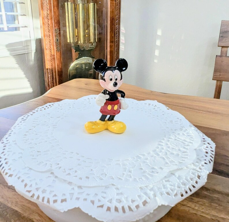 May include: A small Mickey Mouse figurine with black ears, a black body, red shorts, white gloves, and yellow shoes. The figurine is on a white paper doily. The background includes a wooden table and a grandfather clock.