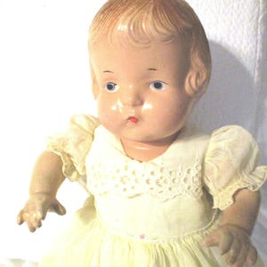 May include: A vintage porcelain doll with blonde hair, blue eyes, and a painted smile. The doll is wearing a white dress with lace trim.