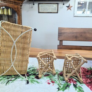 May include: Three woven wicker baskets. One is tall and rectangular with a white metal frame. The other two are smaller, square, and have a crisscross design. All are light brown and sit on a floral tablecloth.