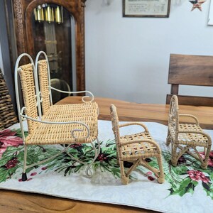 May include: Three miniature wicker chairs. The largest is a rocking chair with a white metal frame. The other two are smaller, with woven seats and backs. The chairs are set on a table runner with a floral pattern.