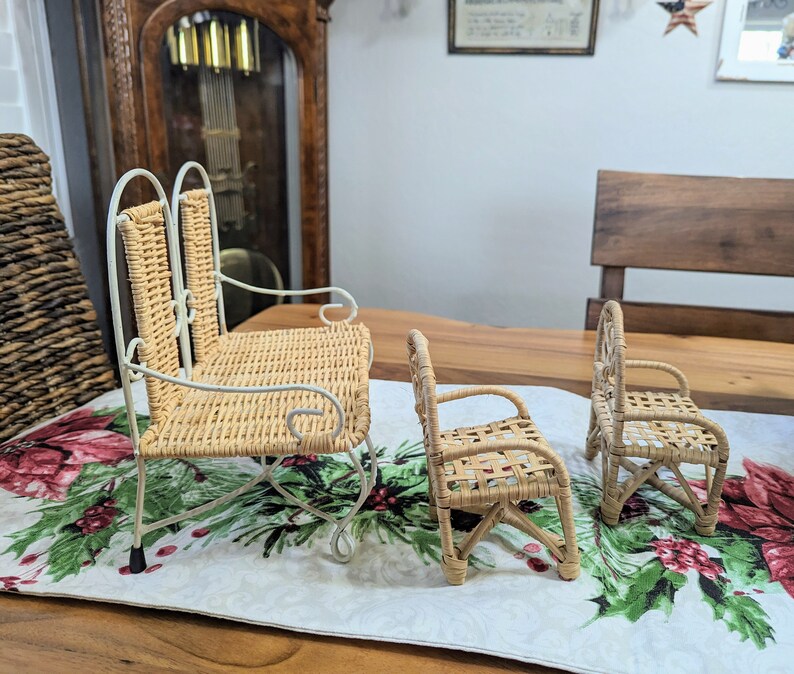 May include: Three miniature wicker chairs. The largest is a rocking chair with a white metal frame. The other two are smaller, with woven seats and backs. The chairs are set on a table runner with a floral pattern.