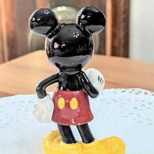 May include: A glossy black Mickey Mouse figurine with large ears, white gloves, a red skirt with yellow buttons, and yellow shoes. The collectible figure is standing on a white doily.