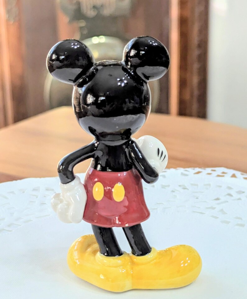 May include: A glossy black Mickey Mouse figurine with large ears, white gloves, a red skirt with yellow buttons, and yellow shoes. The collectible figure is standing on a white doily.