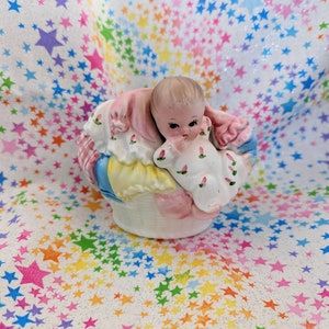 May include: A porcelain figurine of a baby in a pink and blue basket, with a white background featuring a rainbow of stars.