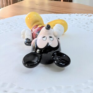 May include: A ceramic Mickey Mouse figurine lying on its back. The figure has a black head and ears, a yellow hat, white gloves, and red shorts. The figurine is on a white, patterned surface.