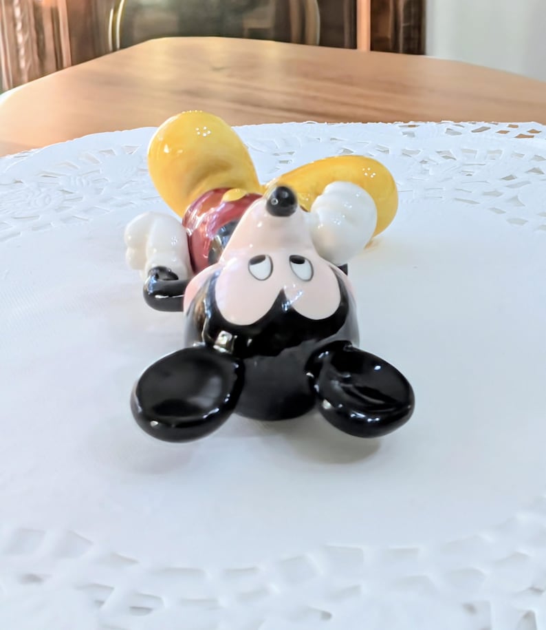 May include: A ceramic Mickey Mouse figurine lying on its back. The figure has a black head and ears, a yellow hat, white gloves, and red shorts. The figurine is on a white, patterned surface.