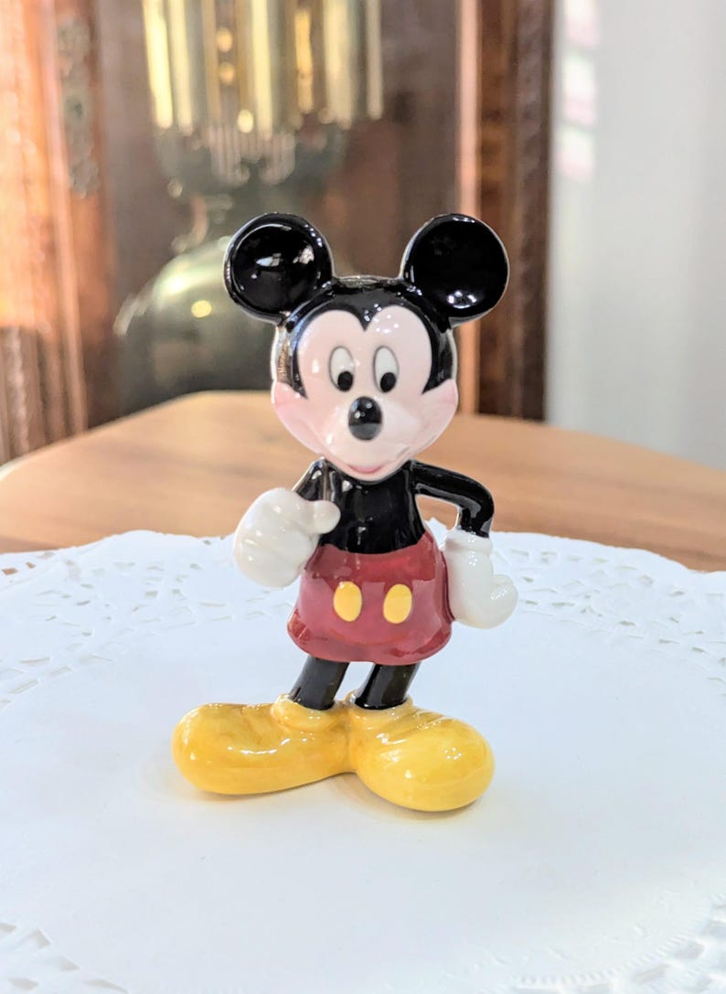 May include: A ceramic Mickey Mouse figurine. The figure has black ears, a black body, red shorts with yellow buttons, white gloves, and yellow shoes. The figurine is standing on a white doily.
