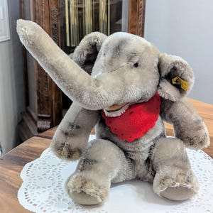 Pode incluir: Um brinquedo de elefante de pel&uacute;cia cinza com uma bandana vermelha. O elefante tem uma tromba longa e est&aacute; sentado em um la&ccedil;o branco.