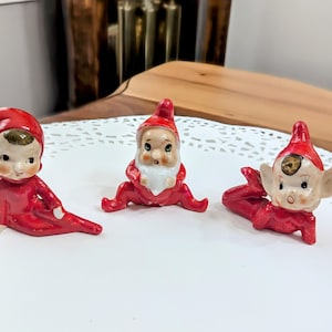 May include: Three vintage ceramic elf figurines in red outfits and hats. The elves have painted faces with rosy cheeks and are in different poses. One is sitting, one is kneeling, and one is lying down.