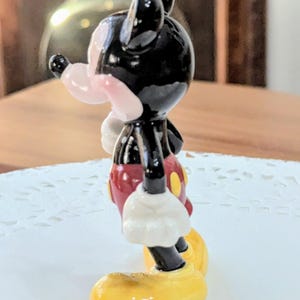 May include: A small figurine of Mickey Mouse, a classic cartoon character. The figure has a black head and ears, a pink nose, and a red shirt with yellow buttons. The figure's gloves are white, and the shoes are yellow.