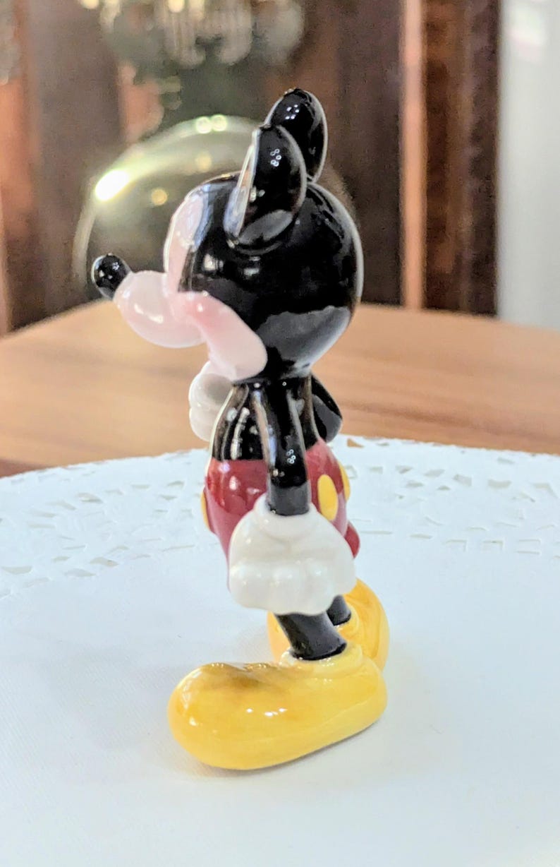 May include: A small figurine of Mickey Mouse, a classic cartoon character. The figure has a black head and ears, a pink nose, and a red shirt with yellow buttons. The figure's gloves are white, and the shoes are yellow.