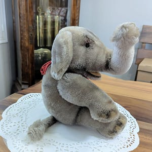 Pode incluir: Um brinquedo de elefante de pel&uacute;cia cinza com uma fita vermelha em volta do pesco&ccedil;o. O elefante est&aacute; sentado em um la&ccedil;o branco.