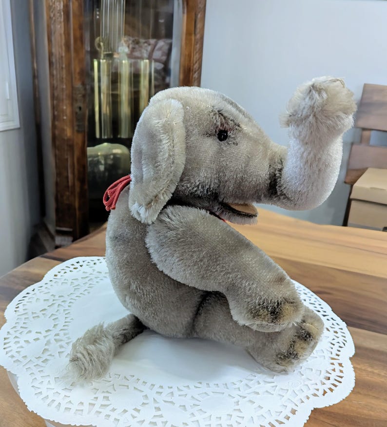 Pode incluir: Um brinquedo de elefante de pel&uacute;cia cinza com uma fita vermelha em volta do pesco&ccedil;o. O elefante est&aacute; sentado em um la&ccedil;o branco.
