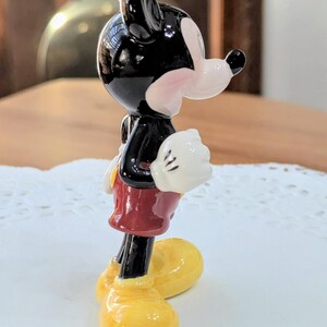 May include: A ceramic Mickey Mouse figurine. The figure has a black head and body, white gloves, red shorts, and yellow shoes. The side view shows the classic cartoon character design. The figurine is a collectible item.