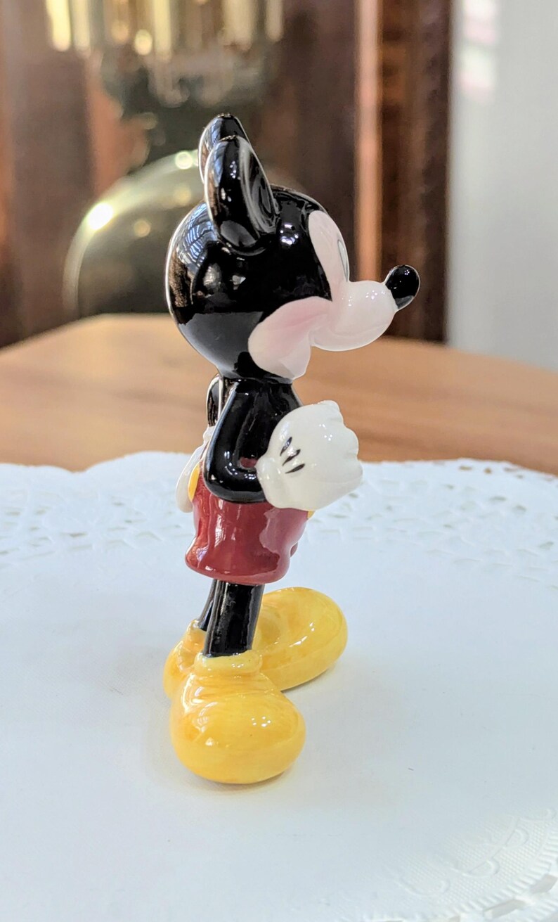 May include: A ceramic Mickey Mouse figurine. The figure has a black head and body, white gloves, red shorts, and yellow shoes. The side view shows the classic cartoon character design. The figurine is a collectible item.