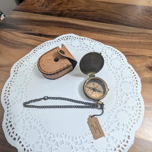 May include: A brass compass with a black needle and a white face with black markings. The compass is attached to a chain and comes with a brown leather case. The case has a flap with a button closure. The compass is on a white doily on a wooden surface. The tag attached to the chain reads "Handmade in Morocco".