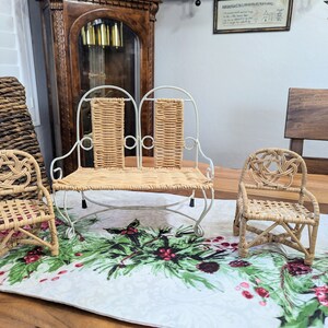May include: Three miniature wicker furniture pieces: a loveseat with a white metal frame and two chairs. The furniture is light brown and sits on a Christmas-themed table runner. A large woven basket and a grandfather clock are in the background.
