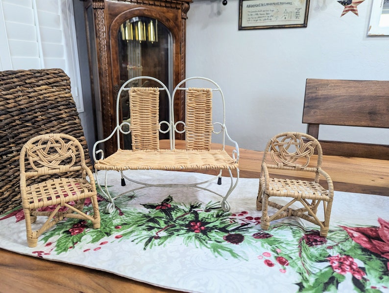 May include: Three miniature wicker furniture pieces: a loveseat with a white metal frame and two chairs. The furniture is light brown and sits on a Christmas-themed table runner. A large woven basket and a grandfather clock are in the background.