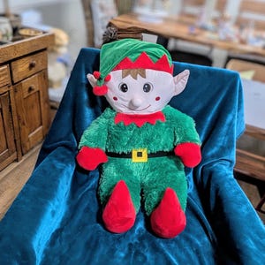 May include: A plush green elf toy with a red hat and a yellow belt. The elf is sitting on a blue velvet chair.
