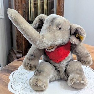Pode incluir: Um brinquedo de elefante de pel&uacute;cia cinza com uma bandana vermelha. O elefante tem uma tromba longa e est&aacute; sentado em um la&ccedil;o branco.