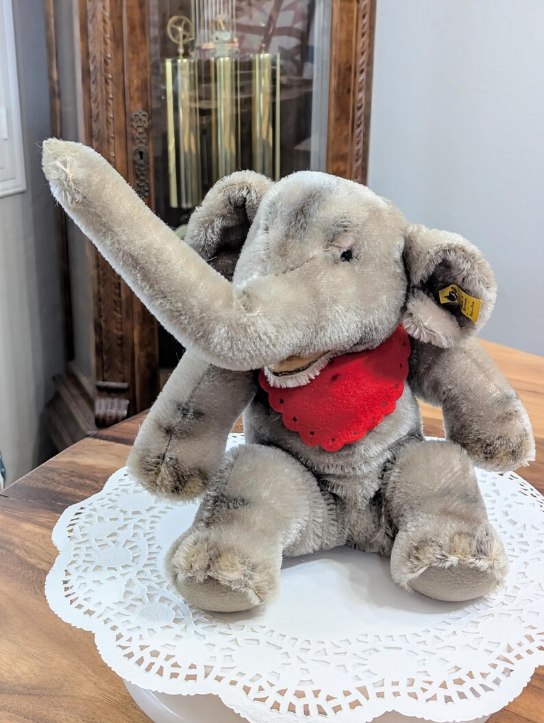 Pode incluir: Um brinquedo de elefante de pel&uacute;cia cinza com uma bandana vermelha. O elefante tem uma tromba longa e est&aacute; sentado em um la&ccedil;o branco.