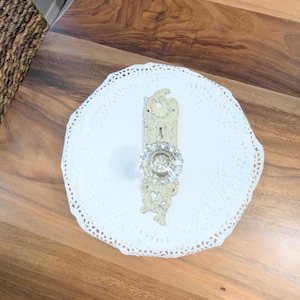 May include: An antique door knob with a clear, faceted glass knob and ornate, aged brass backplate. The door knob is centered on a white, lace-edged doily, suggesting a vintage or decorative theme.