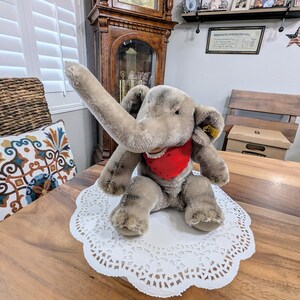 Pode incluir: Um brinquedo de elefante de pel&uacute;cia cinza com uma bandana vermelha sentado em um guardanapo branco em uma mesa de madeira.