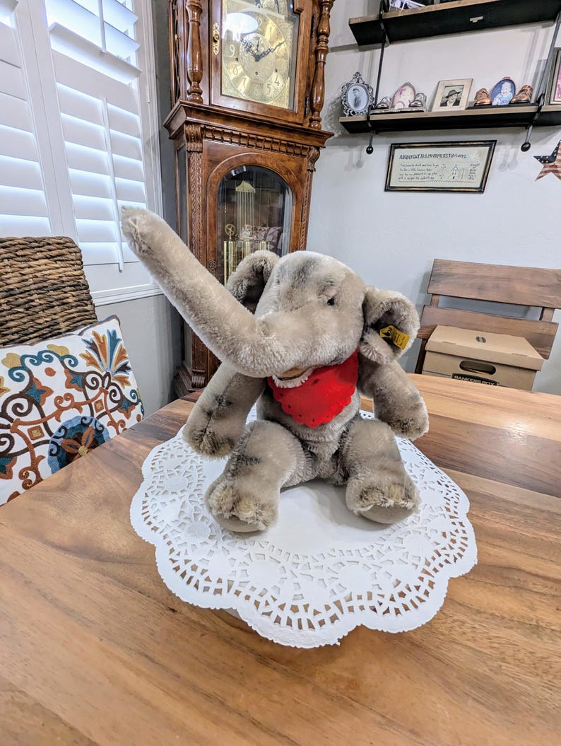 Pode incluir: Um brinquedo de elefante de pel&uacute;cia cinza com uma bandana vermelha sentado em um guardanapo branco em uma mesa de madeira.