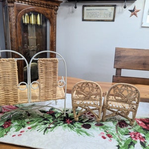 May include: A set of miniature wicker furniture, including a two-seater bench and two armchairs. The furniture has a light brown woven design and white metal frames. The pieces are displayed on a table runner with a festive floral pattern.