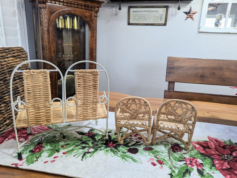 May include: A set of miniature wicker furniture, including a two-seater bench and two armchairs. The furniture has a light brown woven design and white metal frames. The pieces are displayed on a table runner with a festive floral pattern.