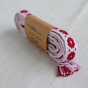 Può includere: Un rotolo di nastro bianco con ricamo floreale rosso. Il nastro è avvolto attorno a un'etichetta di carta marrone che recita "seta - 1 cm x 10 m".