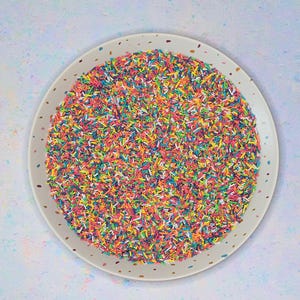 Può includere: Un piatto rotondo e maculato pieno di zuccherini colorati. Gli zuccherini sono un mix di rosso, giallo, verde, blu e arancione, creando un aspetto vibrante e festoso. Il piatto è bianco con macchie marroni.