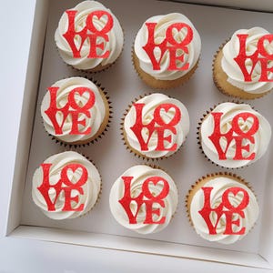 May include: Nine cupcakes in a white box, each topped with white frosting and a red glitter "LOVE" topper. The cupcakes have a light brown base and are arranged in three rows of three.