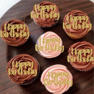 May include: Six cupcakes with chocolate and pink frosting decorated with gold glitter "Happy Birthday" toppers.