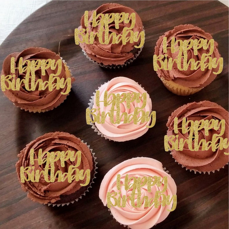 May include: Six cupcakes with chocolate and pink frosting decorated with gold glitter "Happy Birthday" toppers.