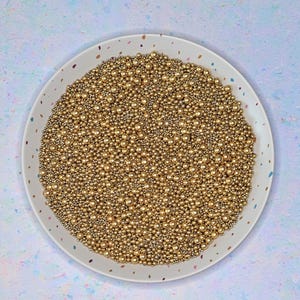 Puede incluir: Un plato blanco lleno de numerosas esferas pequeñas de color dorado. Las esferas varían en tamaño y están densamente empaquetadas, creando una superficie texturizada. El plato tiene un borde moteado y se encuentra sobre una superficie moteada azul claro y blanca.
