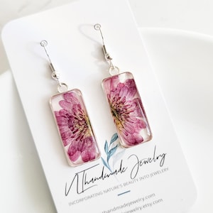 May include: A pair of rectangular earrings featuring pressed pink flowers encased in clear resin. The earrings hang from silver-colored hooks. The earrings are displayed on a white card with the text "Nthhandmade Jewelry".