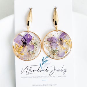 May include: A pair of gold-toned earrings with a circular pendant design. Each pendant features a clear resin encasing purple and yellow dried flowers and gold flakes. The earrings are displayed on a white card with the text "Nthhandmade Jewelry".