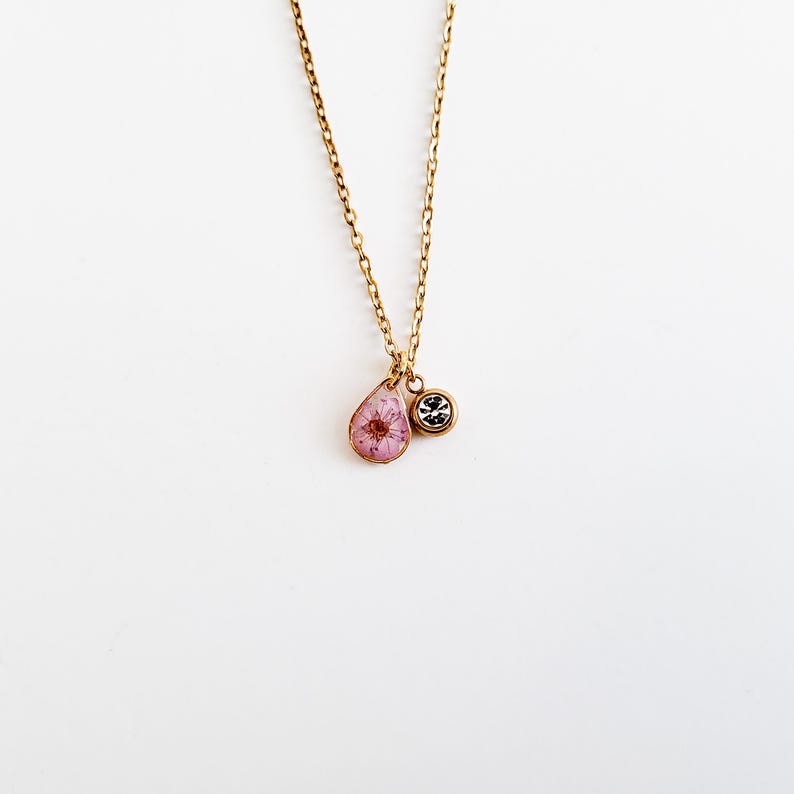 Op de afbeelding: Een goudkleurige ketting met twee hangers. E&eacute;n hanger is een druppelvorm met een roze bloem in heldere hars. De andere hanger is een kleine, ronde bedel met een zwart-zilveren ontwerp. De ketting hangt tegen een witte achtergrond.