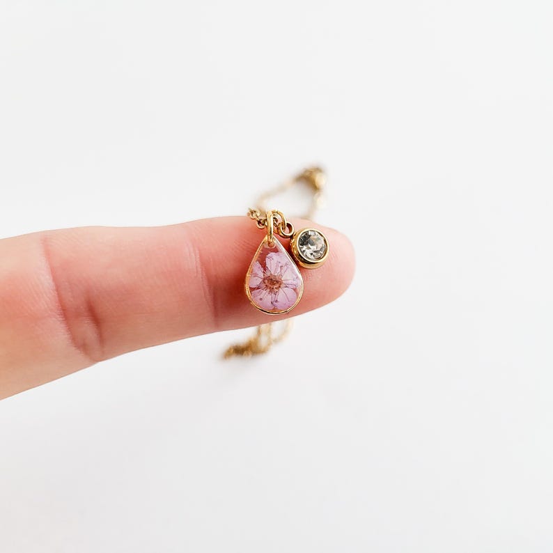 Op de afbeelding: Een goudkleurige ketting met een hanger met een roze bloem in heldere hars. Een kleine, ronde, heldere edelsteenbedel hangt naast de hanger. De ketting wordt getoond tegen een witte achtergrond.