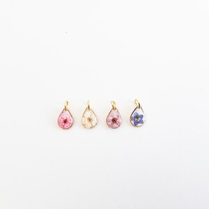 Op de afbeelding: Vier druppelvormige hangers met goudkleurige frames, elk met een andere gedroogde bloem. De bloemen zijn roze, wit, roze met rood en blauw. De hangers worden op een witte achtergrond weergegeven.