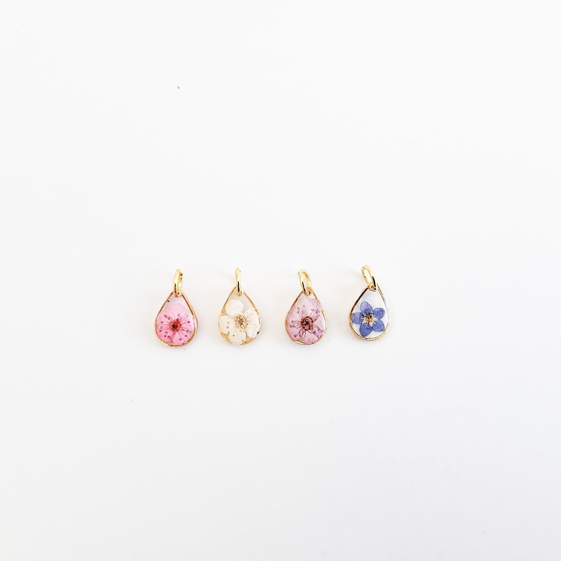 Op de afbeelding: Vier druppelvormige hangers met goudkleurige frames, elk met een andere gedroogde bloem. De bloemen zijn roze, wit, roze met rood en blauw. De hangers worden op een witte achtergrond weergegeven.