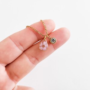 Op de afbeelding: Een goudkleurige ketting met een druppelvormige hanger met een geperste roze bloem in heldere hars. Een kleine, ronde, heldere kristallen bedel hangt naast de hanger. De ketting wordt getoond tegen een lichte achtergrond.