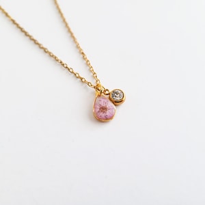 Op de afbeelding: Een goudkleurige ketting met een fijne ketting. De hanger heeft een druppelvormige bedel met een roze bloem in hars en een kleine, ronde, heldere edelsteen. De ketting wordt gepresenteerd tegen een witte achtergrond.