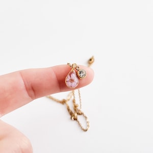 Op de afbeelding: Een goudkleurige ketting met een druppelvormige hanger met een kleine roze bloem. Een kleine, ronde, heldere edelsteen is aan de hanger bevestigd. De ketting wordt gepresenteerd tegen een witte achtergrond.