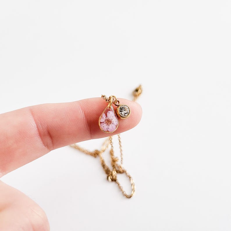 Op de afbeelding: Een goudkleurige ketting met een druppelvormige hanger met een kleine roze bloem. Een kleine, ronde, heldere edelsteen is aan de hanger bevestigd. De ketting wordt gepresenteerd tegen een witte achtergrond.