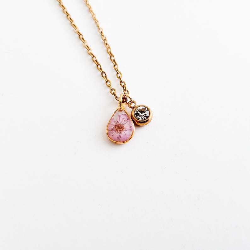 Op de afbeelding: Een goudkleurige ketting met twee hangers. E&eacute;n hanger is een druppelvorm met een roze bloem erin. De andere hanger is een kleine, ronde, heldere edelsteen. De ketting is ook goudkleurig.