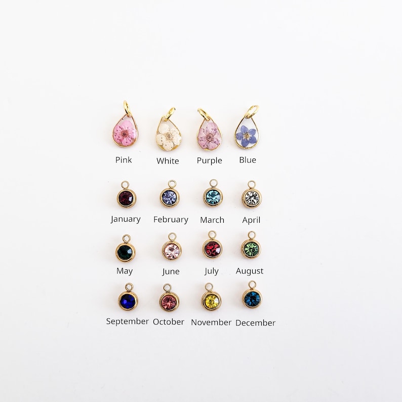 Op de afbeelding: Een verzameling goudkleurige hangers met gekleurde edelstenen en bloemen. De hangers zijn gelabeld met maanden en kleuren, waaronder roze, wit, paars en blauw. De afbeelding staat op een witte achtergrond.