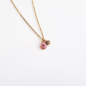 Op de afbeelding: Een delicate goudkleurige ketting met een druppelvormige hanger met een roze bloem. Een kleine, ronde, heldere edelsteen hangt naast de hanger. De ketting staat op een witte achtergrond.